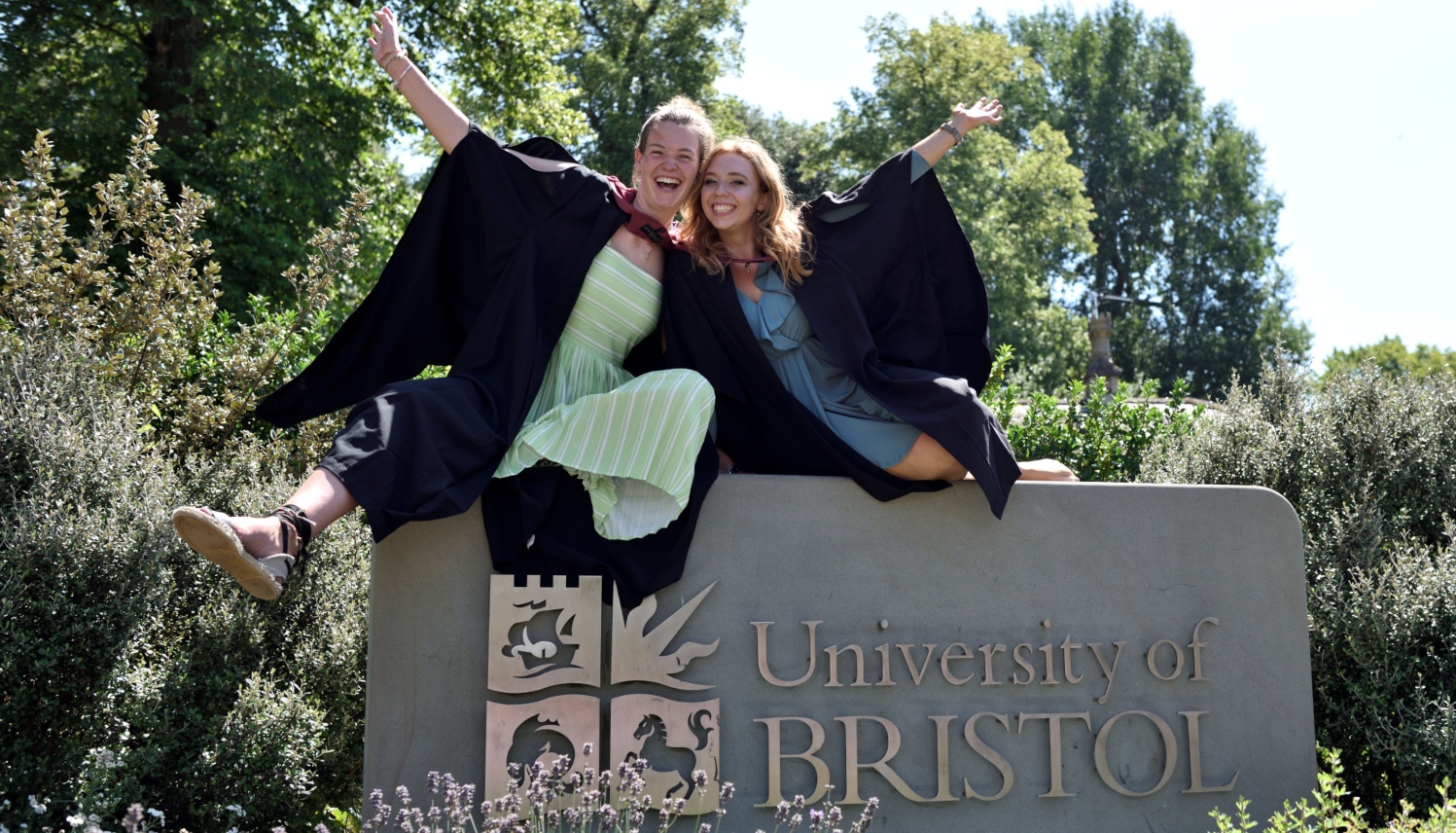 Two students in Degree gowns sitting on a University of Bristol sign.